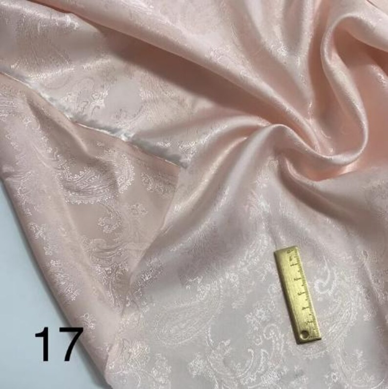May include: A pale pink fabric with a white paisley pattern. The fabric is smooth and silky. A ruler is included for scale.