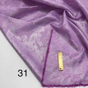 May include: A close-up of a purple fabric with a white paisley pattern. The fabric has a soft, silky texture and a subtle sheen. The fabric is folded in half and the edge is trimmed with a pink fringe. The number "31" is written on the fabric.