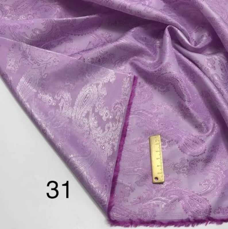 May include: A close-up of a purple fabric with a white paisley pattern. The fabric has a soft, silky texture and a subtle sheen. The fabric is folded in half and the edge is trimmed with a pink fringe. The number "31" is written on the fabric.