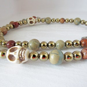 May include: A beaded necklace with brown, beige, and green stones, and two white skull beads. The necklace has a silver clasp.
