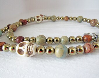 May include: A beaded necklace with brown, beige, and green stones, and two white skull beads. The necklace has a silver clasp.