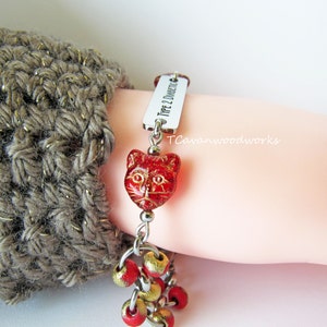 May include: A silver bracelet with a red cat charm and a silver tag that reads "Type 2 Diabetic". The bracelet also has red and gold beads.