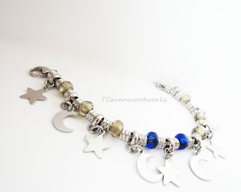 May include: A silver charm bracelet with silver stars, moons, and blue glass beads. The bracelet has a clasp closure and the text "TCavanwoodworks" is visible on the bracelet.