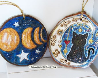 May include: Two round wood slice ornaments with a blue background and gold glitter. One ornament features a full moon and crescent moons with white stars. The other ornament features a white crescent moon with a face and a black cat silhouette.