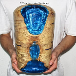 May include: A cylindrical wooden vase with a natural wood grain finish. The vase features two sections of vibrant blue resin, creating a striking contrast. The text "TCavanwoodworks" is visible at the top.