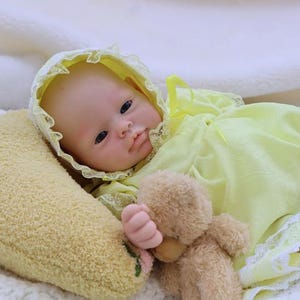 May include: A lifelike baby doll wearing a yellow dress and bonnet with lace trim, lying on a soft, yellow pillow. The doll is holding a small, brown teddy bear. The background is a white, textured blanket.