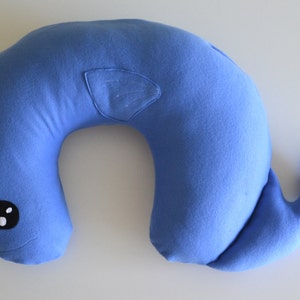 Puede incluir: Una almohada de viaje azul en forma de narval con un cuerno blanco y un ojo negro. La almohada está diseñada para sostener el cuello y la cabeza.