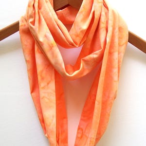 May include: An orange infinity scarf with a tie-dye pattern hangs on a wooden hanger. The scarf is made of soft fabric and has a seamless, circular design. The color is a blend of orange and peach.