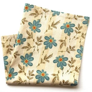 May include: Folded cream-colored fabric with a repeating floral pattern. The design features blue flowers with orange centers and brown leafy sprigs against a striped background. The fabric is folded, showing the pattern on both sides.