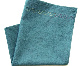 May include: Teal-colored linen napkin with a decorative, wavy stitch in yellow and purple thread along one edge. The napkin is folded, showcasing the texture of the fabric. Ideal for kitchen or dining decor.