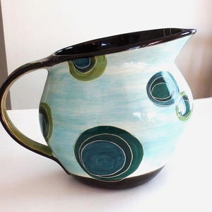 May include: A ceramic pitcher featuring a light blue base with hand-painted dark green and teal concentric circle patterns. The handle, rim, and base are finished in a solid dark brown glaze, providing a contrast to the light blue body.