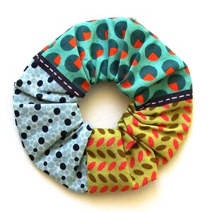 May include: A colorful scrunchie with a variety of patterns. The scrunchie features sections of teal fabric with orange and teal circles, light blue fabric with dark blue polka dots, and olive green fabric with red and brown ovals.