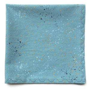 May include: A square, teal-colored cloth napkin with a scalloped edge. The fabric is speckled with white, gold, and navy blue paint-like splatters, creating a textured appearance. The napkin is likely made of cotton or a similar fabric.