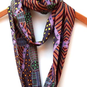 May include: An infinity scarf featuring a complex pattern of dots, stripes, and geometric shapes in shades of dark brown, orange, purple, green, and yellow. The fabric is draped in a continuous loop, displaying a dense, multi-colored design.