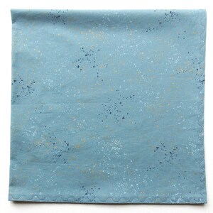 May include: A square, light blue fabric with a speckled pattern in white, navy, and gold. The edges are finished with a scalloped stitch. This fabric could be used for quilting or other craft projects.