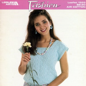 May include: A woman wearing a light blue knitted top with a square neckline and short sleeves. She is holding a white carnation and wearing a multi-colored beaded necklace. The top is paired with a white skirt and a floral patterned scarf tied around her waist. The image is from a knitting pattern booklet featuring "Nomotta Air Cotton".