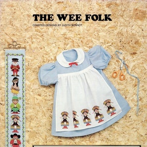 May include: A cross-stitch pattern for a sampler featuring a variety of characters in traditional clothing. The pattern is titled "The Wee Folk" and includes a variety of characters, including a leprechaun, a Native American, a Scottish Highlander, and a Dutch girl. The pattern is charted by Judith Bonnot.