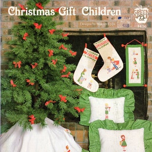 May include: A Christmas tree skirt with a green ruffle and white background featuring embroidered children holding gifts. Two stockings with embroidered children and names "Bryan" and "Merrimon" hang above a fireplace. Two pillows with embroidered children are on the floor. The text "Christmas Gift Children" is at the top of the image. The text "Book #522" is at the bottom of the image.