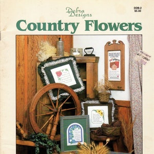 May include: A collection of cross stitch patterns featuring country flowers, a spinning wheel, a basket of sunflowers, and a vase of red flowers. The patterns are framed and displayed in a rustic setting. The title "Country Flowers" is printed at the top of the page. The text "Designs in Counted Cross Stitch" is printed at the bottom of the page.