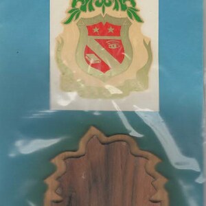 May include: A wooden paddle-shaped item with a decorative emblem and a separate wooden piece. The emblem features a shield design with green, red, and gold accents. The text "THE PADDLE TRAMPS MFG. CO." is visible.
