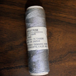 May include: A spool of silver lame thread with a white label that reads "WESTRIM PLY LAME 10649 LENGTH: 75 YARDS MADE IN JAPAN ESTERN TRIMMING CORP. ATSWORTH, CALIF. 91311"