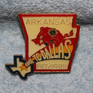 May include: A gold-colored pin with a red and white outline of the state of Arkansas. The pin features a red cartoon wild boar with a blue and yellow banner that reads "Back to Dallas 1917/1985".