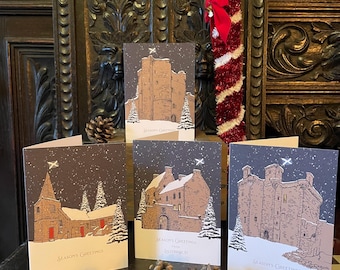 May include: Set of five Christmas greeting cards featuring illustrations of Scottish castles and buildings in winter. Each card has a snowy scene with the text "Season's Greetings". Pine cones are arranged in front of the cards.
