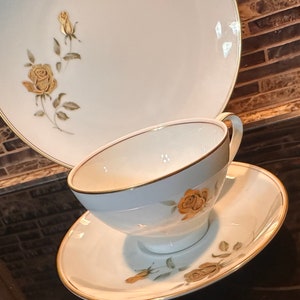 May include: White porcelain teacup, saucer, and plate set with gold trim and a floral rose design. The teacup and saucer are in the foreground, with the plate in the background. The roses are a muted gold color.