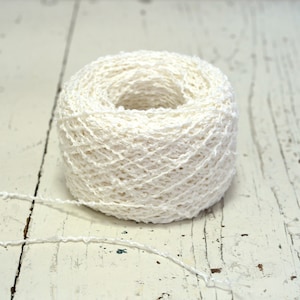 Puede incluir: Una madeja de hilo blanco roto con un tejido texturizado y abierto. El hilo es cilíndrico con un agujero en el centro. Un hilo se extiende desde la madeja, descansando sobre una superficie de madera blanca desgastada.