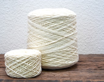 May include: Two spools of off-white yarn sit on a wooden surface. The larger spool is cylindrical, and the smaller one is a shorter, wider cylinder. The yarn is tightly wound, with visible crisscrossing threads. The background is a textured white wall.