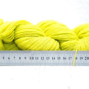 Pode incluir: Um novelo de fio amarelo brilhante medido com uma r&eacute;gua. A r&eacute;gua mostra medidas em polegadas e cent&iacute;metros.