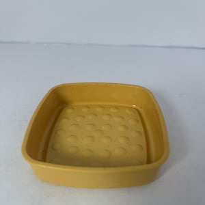 May include: A small, square, yellow plastic tray with rounded corners. The interior base features a pattern of evenly spaced, raised circular bumps. The tray's sides are slightly sloped, and the overall design is simple and functional.
