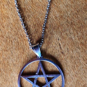 Puede incluir: Un collar de metal plateado que presenta un colgante circular con un diseño de pentagrama recortado. El colgante está unido a una cadena de eslabones simple que incluye un cierre de mosquetón en la parte superior, con un acabado metálico pulido.