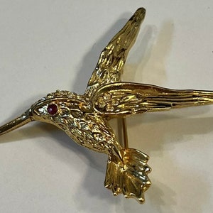 May include: A gold-toned hummingbird brooch with a red rhinestone eye and clear rhinestone accents.