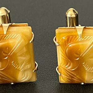 May include: A pair of gold cufflinks with a carved amber design. The design features a man's face with a helmet.