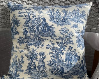 May include: Square throw pillow with a blue and white toile pattern. The design features pastoral scenes with figures, trees, and buildings. The pillow is placed on a white wicker chair.