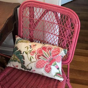May include: A pink wicker chair with a floral patterned pillow. The pillow features a large red and pink flower design with green leaves on a cream background. The chair is a vibrant shade of pink.