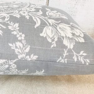 May include: Gray decorative pillow with a white floral pattern. The pillow features a detailed design of flowers, leaves, and bows. The pillow has a zipper closure and is made of a textured fabric.