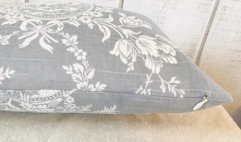 May include: Gray decorative pillow with a white floral pattern. The pillow features a detailed design of flowers, leaves, and bows. The pillow has a zipper closure and is made of a textured fabric.