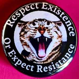 May include: Round black button with the text "Respect Existence Or Expect Resistance" in white. The button features a close-up image of a snarling cat with its mouth open, showing its teeth and pink tongue.