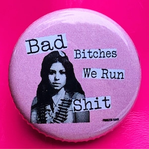 May include: A round, pink button with the text "Bad Bitches We Run Shit" in a bold, black font. The button features a black and white image of a woman. The text "Princess Radmi" is at the bottom.