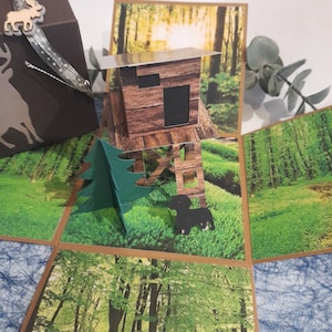 Könnte beinhalten: Eine handgefertigte braune Geschenkbox aus Papier mit einem Elch aus Holz und einem silbernen Band. Die Box öffnet sich und zeigt eine Pop-up-Waldszene mit einer Hütte, einem grünen Baum und einem schwarzen Hund. Der Hintergrund zeigt eine Waldlandschaft.