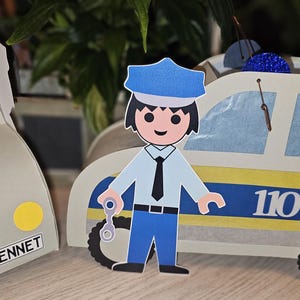 Könnte beinhalten: Eine handgefertigte Papierbastelarbeit mit einer Polizistenfigur und zwei Spielzeugautos. Das Polizeiauto hat blaue und gelbe Streifen, und die Nummer "110" ist sichtbar. Ein Auto hat den Namen "BENNET" auf dem Nummernschild.