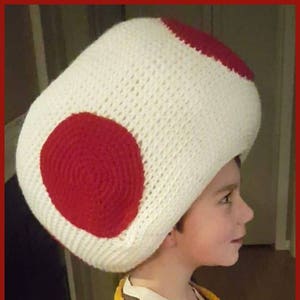 May include: A child wearing a white and red crocheted mushroom hat. The hat has a large red circle on the side. The child is wearing a blue and yellow crocheted vest.