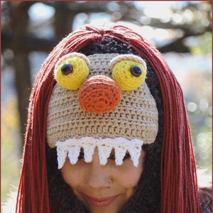 May include: A crocheted hat with a red yarn wig, large yellow eyes, an orange nose, and white teeth. The hat is being worn by a person with a brown fur scarf and a silver necklace.