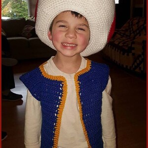 May include: A young child wearing a blue and yellow crocheted vest and a white crocheted mushroom hat with a red top. The child is smiling and looking at the camera.