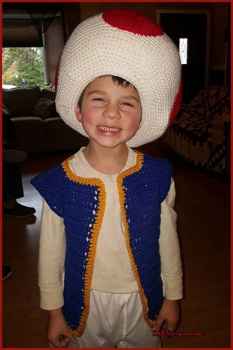 May include: A young child wearing a blue and yellow crocheted vest and a white crocheted mushroom hat with a red top. The child is smiling and looking at the camera.