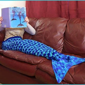 May include: A blue and turquoise crocheted mermaid tail blanket. The blanket is draped over a brown leather couch. The person is holding a book with the title "Fantastic Journeys" on the cover.