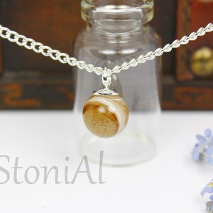 Puede incluir: Collar de cadena plateada con un colgante esf&eacute;rico con un dise&ntilde;o veteado en marr&oacute;n claro y blanco. El collar se muestra frente a una botella de vidrio y un fondo de madera. El texto "StoniAl" es visible.