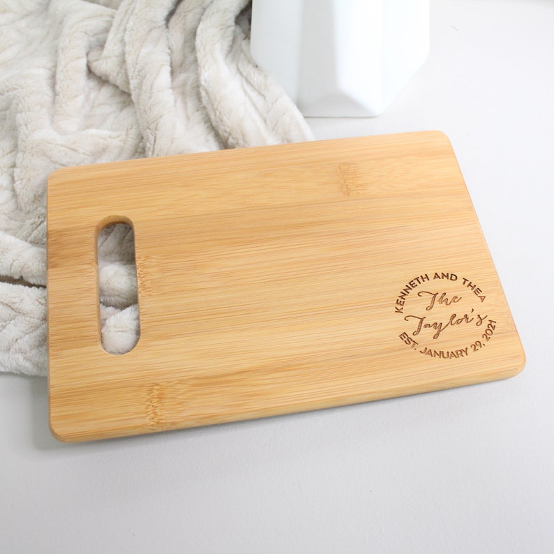 May include: A natural wood cutting board with a handle. The board is engraved with the text "Kenneth and Thea The Taylor's Est. January 29, 2021".